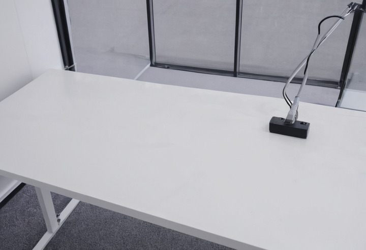 A white office desk seen from above with a lux meter sensor lying on the desk surface under a linear ceiling luminaire.