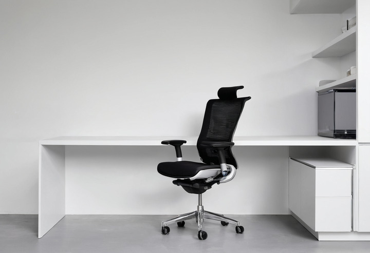 A black mesh office task chair with adjustable armrests standing beside a white kitchen wall cupboard in a white studio.