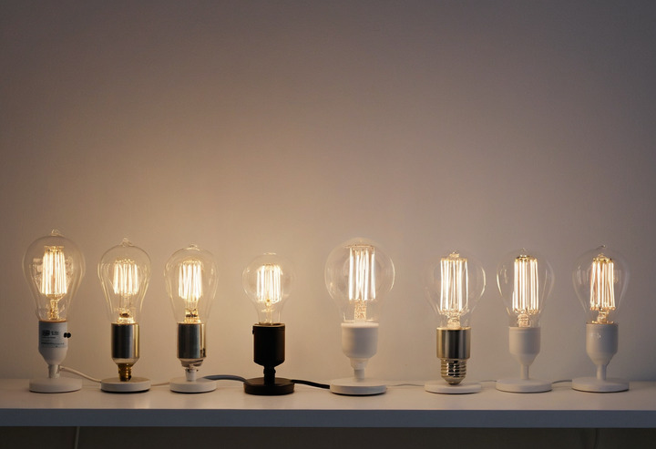 Five bare lamps in a row on a white shelf: a halogen bulb, a compact fluorescent, a filament LED, a frosted LED and a linear LED tube.