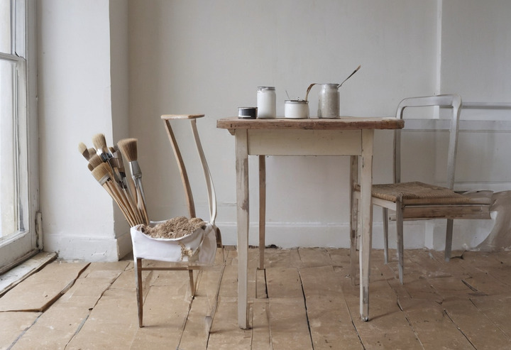 A pale wooden dining chair and an open tin of matt white paint with a brush on a paper-covered table in a white room.