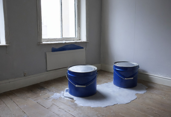 A half-painted white wall with a roller tray and two tins of paint on a dust sheet in front of it.